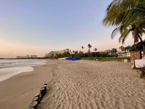 A beach with a sandy shore, palm trees, and a building in the background. - Vista Pacífico • Beachfront Condo • Pool • Ixtapa-Zihuatanejo (Ixtapa Zihuatanejo)