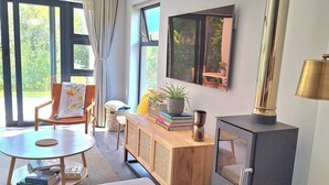 A living room with a sofa, a table, a wooden cabinet, a large window, and a wood-burning stove. - Rustling Reeds Sanctuary (Cape Town)
