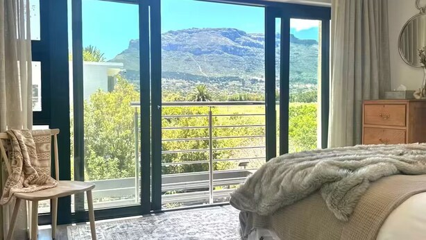 A bedroom with a large bed, a chair, a wooden dresser, and a view of a mountainous landscape through a sliding glass door. - Rustling Reeds Sanctuary (Cape Town)