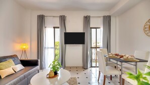 Ein modernes Wohnzimmer mit einer Couch, einem Esstisch, Stühlen, einem Flachbildfernseher und einem großen Fenster mit Vorhängen.