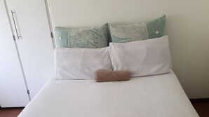 Una cama bien tendida con ropa de cama blanca y una toalla marrón, dos almohadas con texto, y un armario blanco a la izquierda.