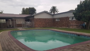 Una piscina rectangular con borde de ladrillo, una casa de fachada de ladrillo y una palmera al fondo.
