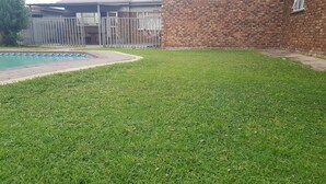 Un patio trasero con césped, una piscina, un muro de ladrillo y una cerca de metal.