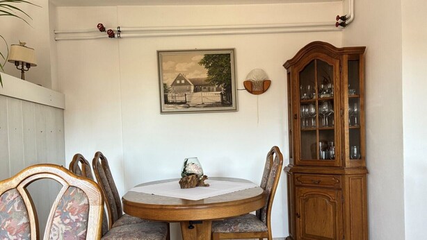 Un comedor con una mesa redonda de madera, seis sillas, un aparador con cristalería y un cuadro en la pared.