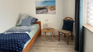 Un dormitorio pequeño y ordenado con cama, una silla, una mesita y una ventana con cortinas.