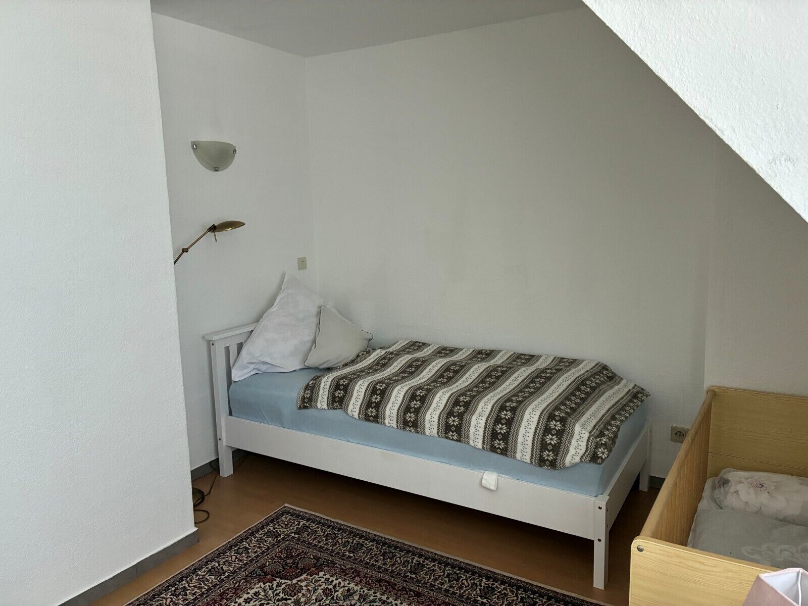 Habitació petita amb un llit individual, sostre inclinat, estenida amb un sobrellit estampat, una làmpada de nit i una catifa al terra.
