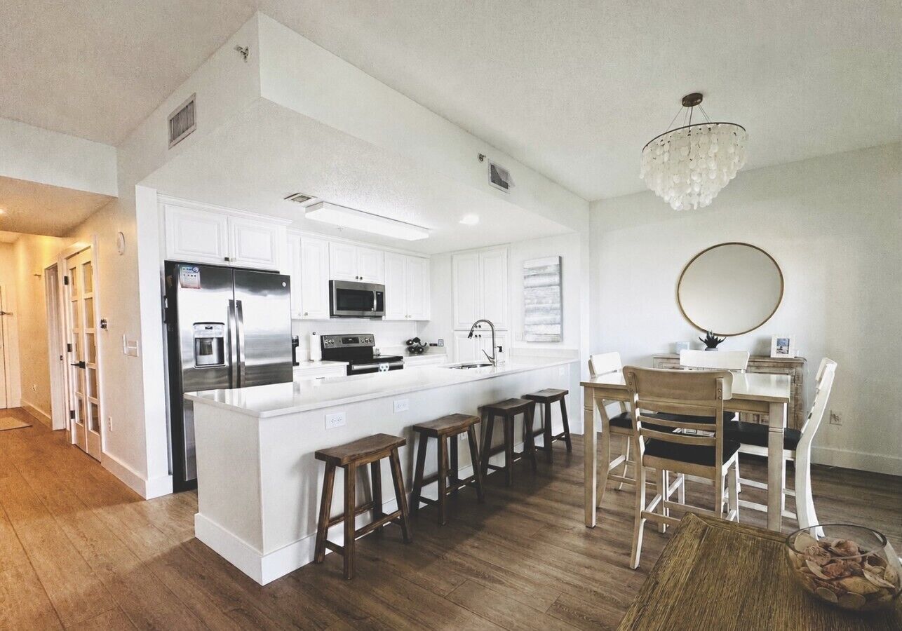 A modern kitchen with white cabinetry, a central island with stools, a dining area with wooden chairs, and a chandelier.