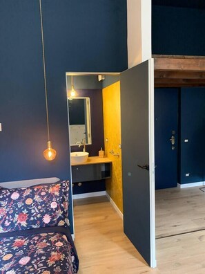 Ein Schlafzimmer mit einem Bett, einer Nachttischlampe und Blick in ein Badezimmer mit Waschbecken und Spiegel.