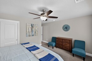 Ein Schlafzimmer mit Bett, Deckenventilator, Kommode, zwei Stühlen und einer Wanddekoration.