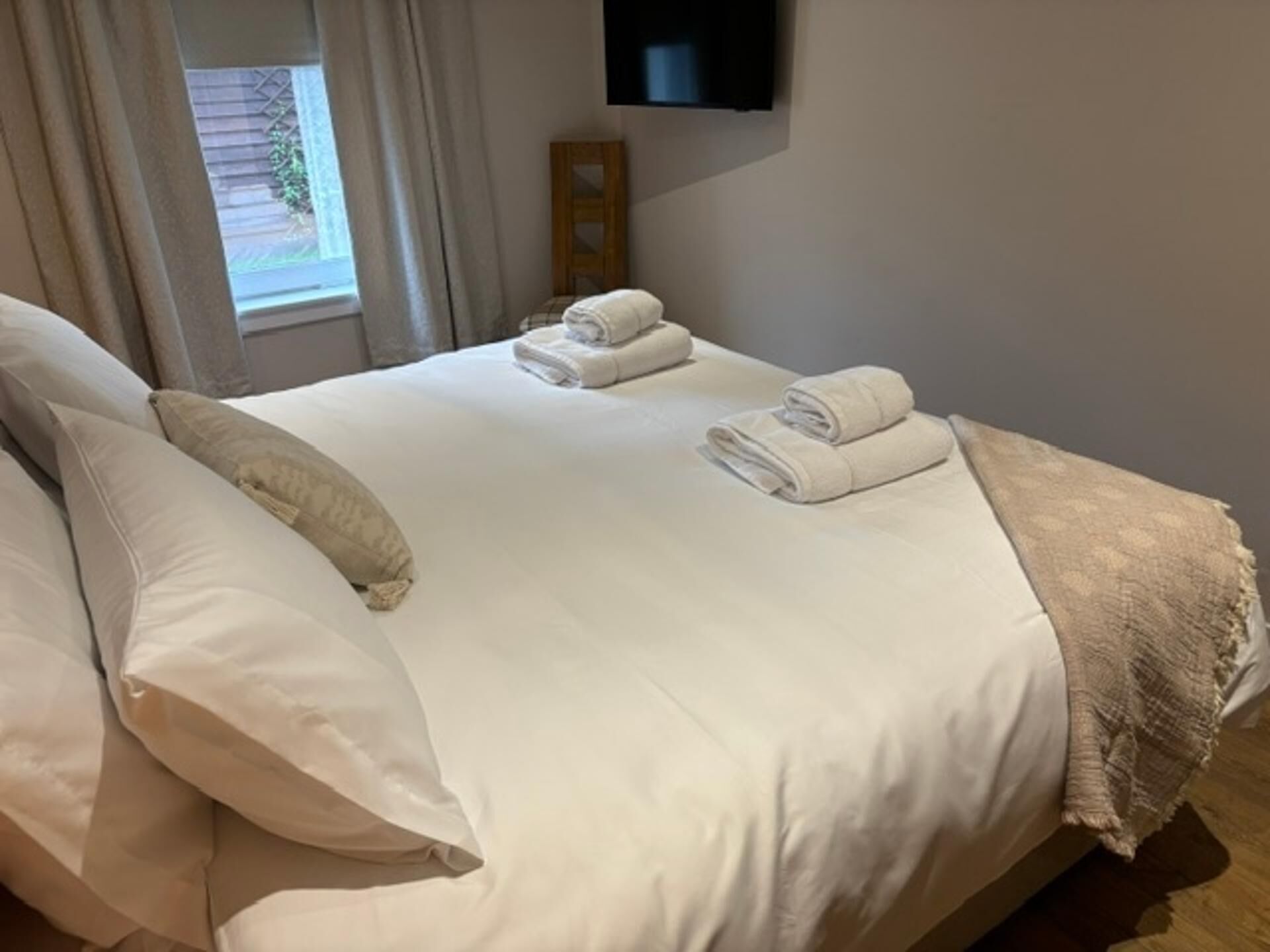 A neatly made bed with pillows and folded towels, a wooden headboard, a television mounted on the wall, and a window with curtains.