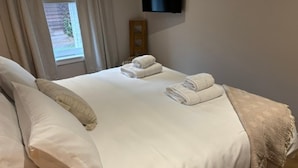 A neatly made bed with pillows and folded towels, a wooden headboard, a television mounted on the wall, and a window with curtains. - Cosy two Bedroom Bungalow With Garden in Ballater (Ballater)