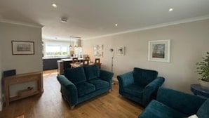 A living room with a sofa, armchairs, and a coffee table. There is a dining area in the background with a table and chairs. - Cosy two Bedroom Bungalow With Garden in Ballater (Ballater)