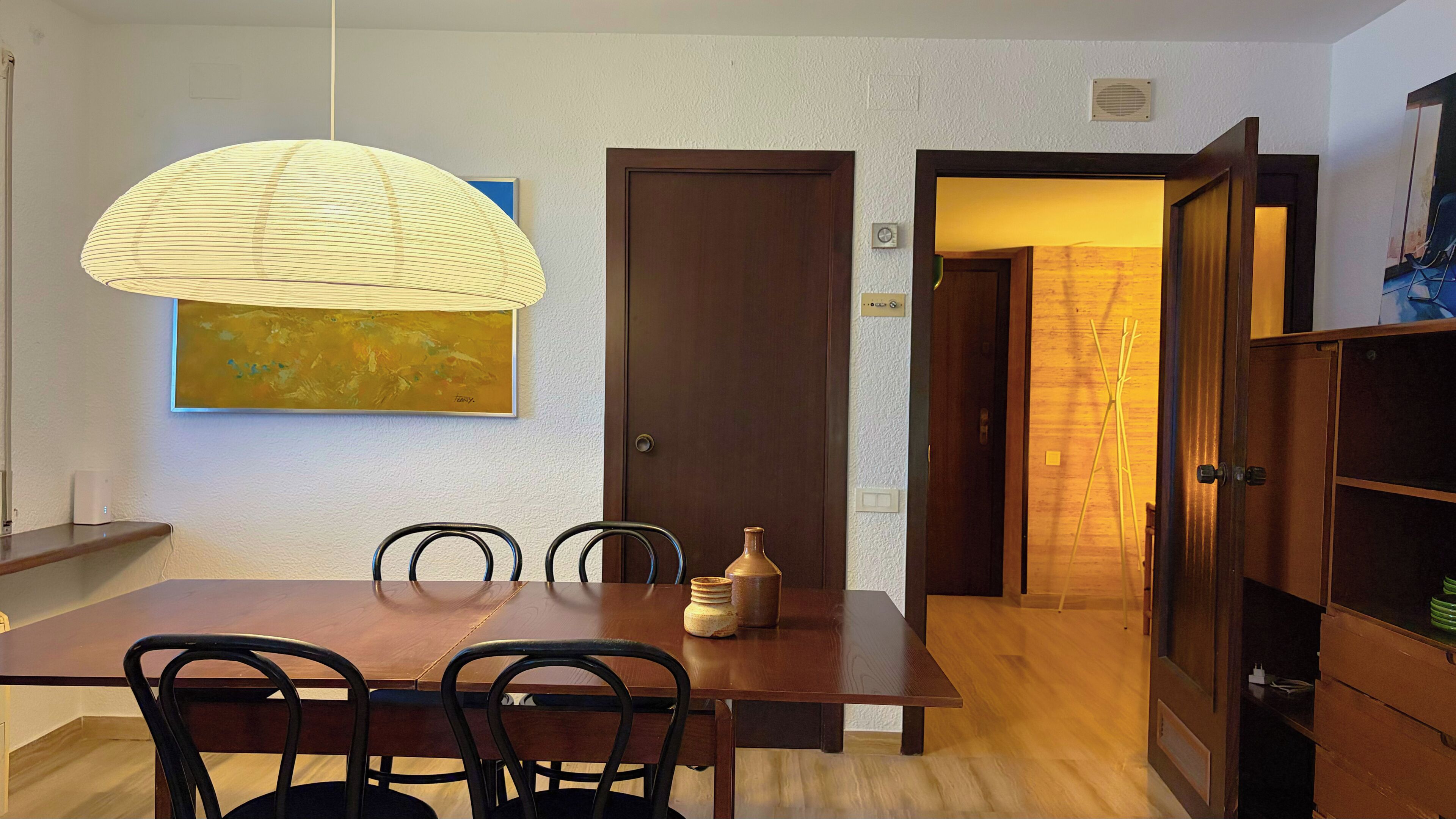 Een eetruimte met een houten tafel, stoelen, een hangende lamp, een deur en een schilderij aan de muur.