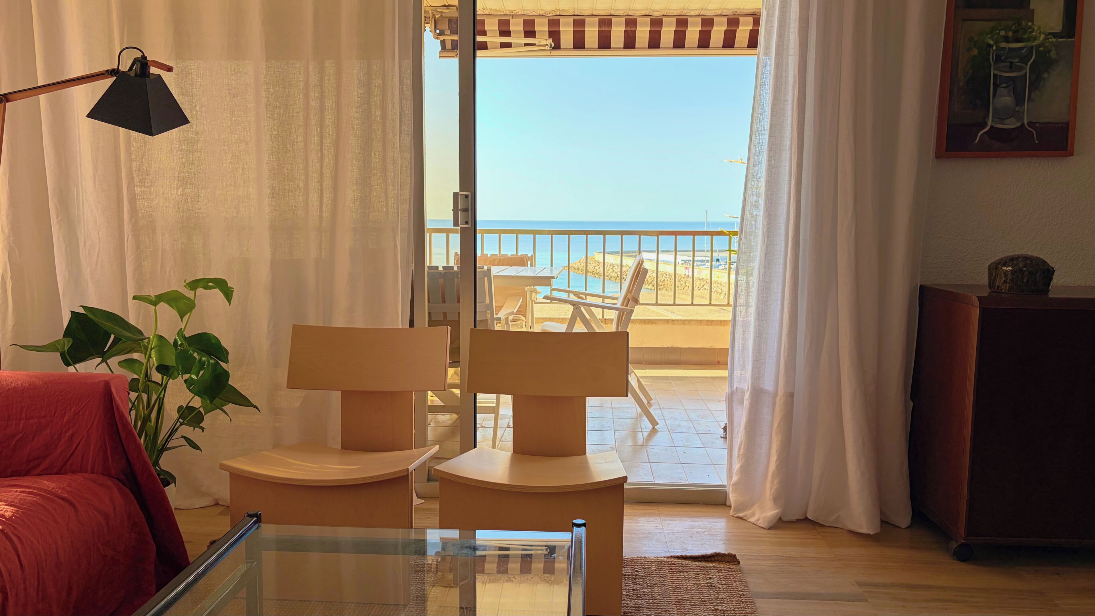 Una habitación con una mesa de vidrio, dos sillas, un sofá rojo, una lámpara y vistas a la playa.