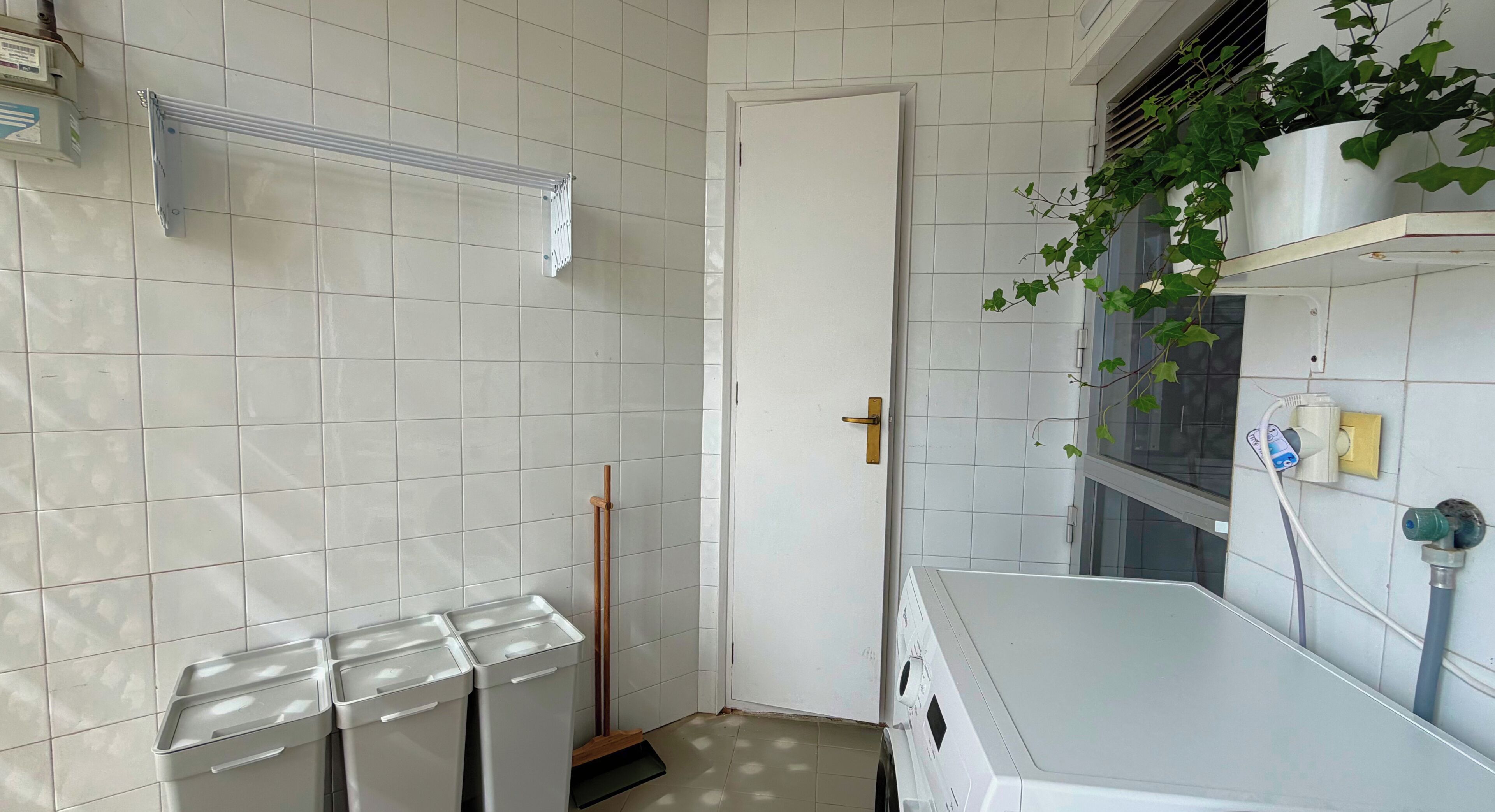 A small, clean room with white tiled walls, a door, a radiator, a mop, a washing machine, and a potted plant.