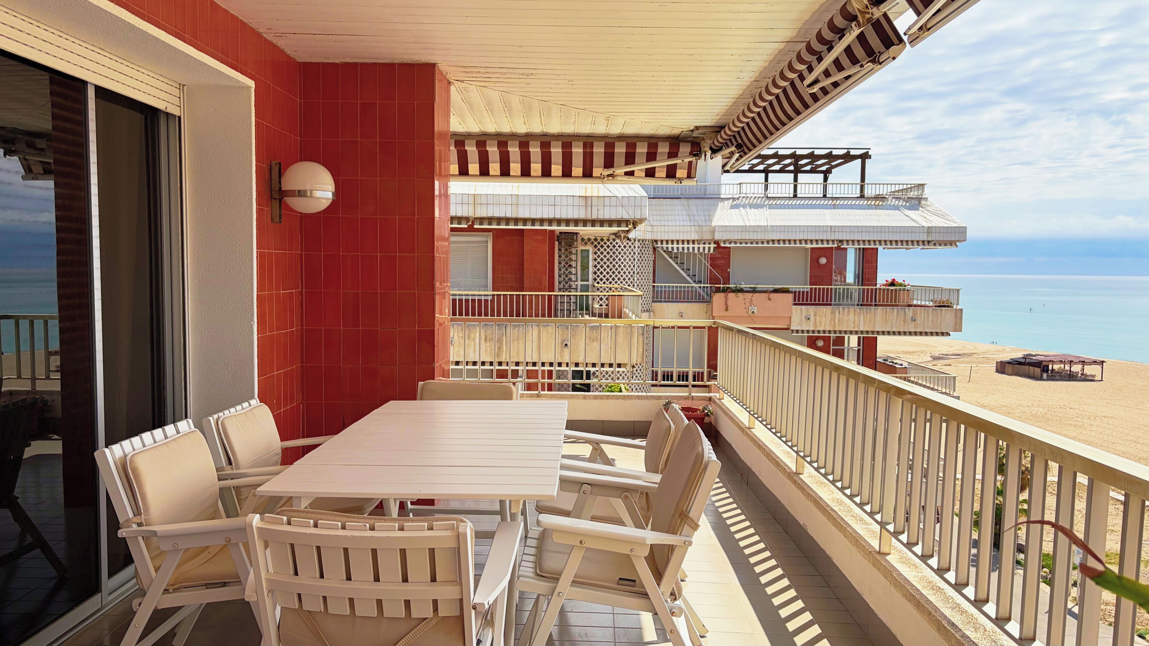 A balcony with a table and chairs, a view of the sea, and a building with multiple balconies.