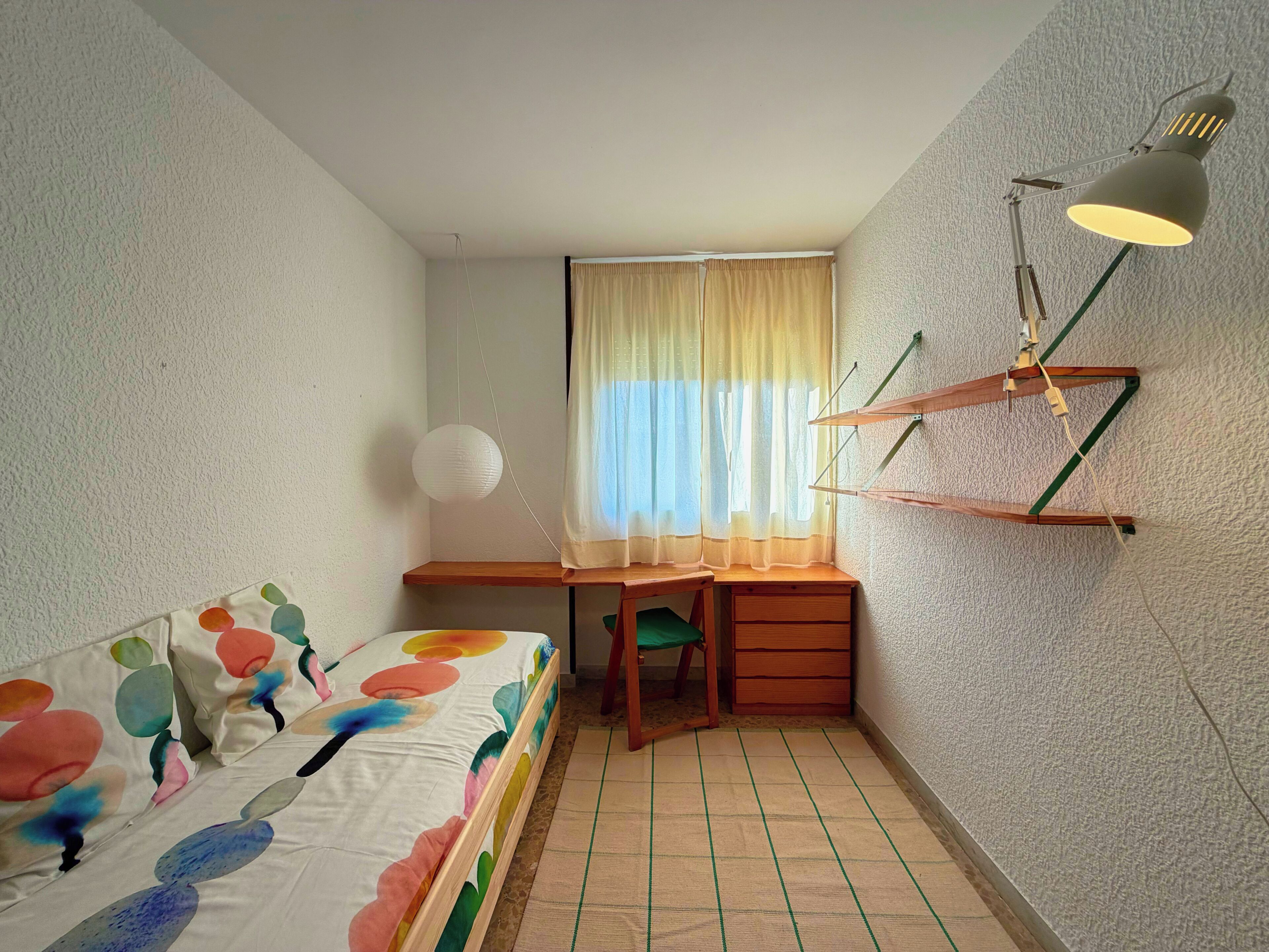 A small, compact room with a bed, a desk, a chair, a hanging lamp, and a window with curtains.