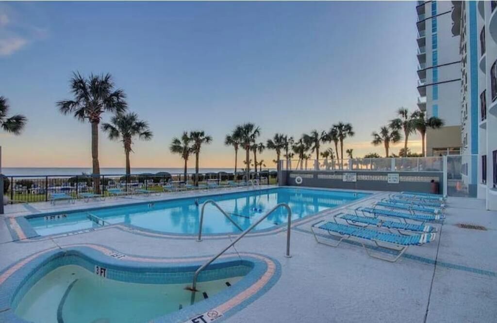 A pool area with lounge chairs, palm trees, and a building with balconies.