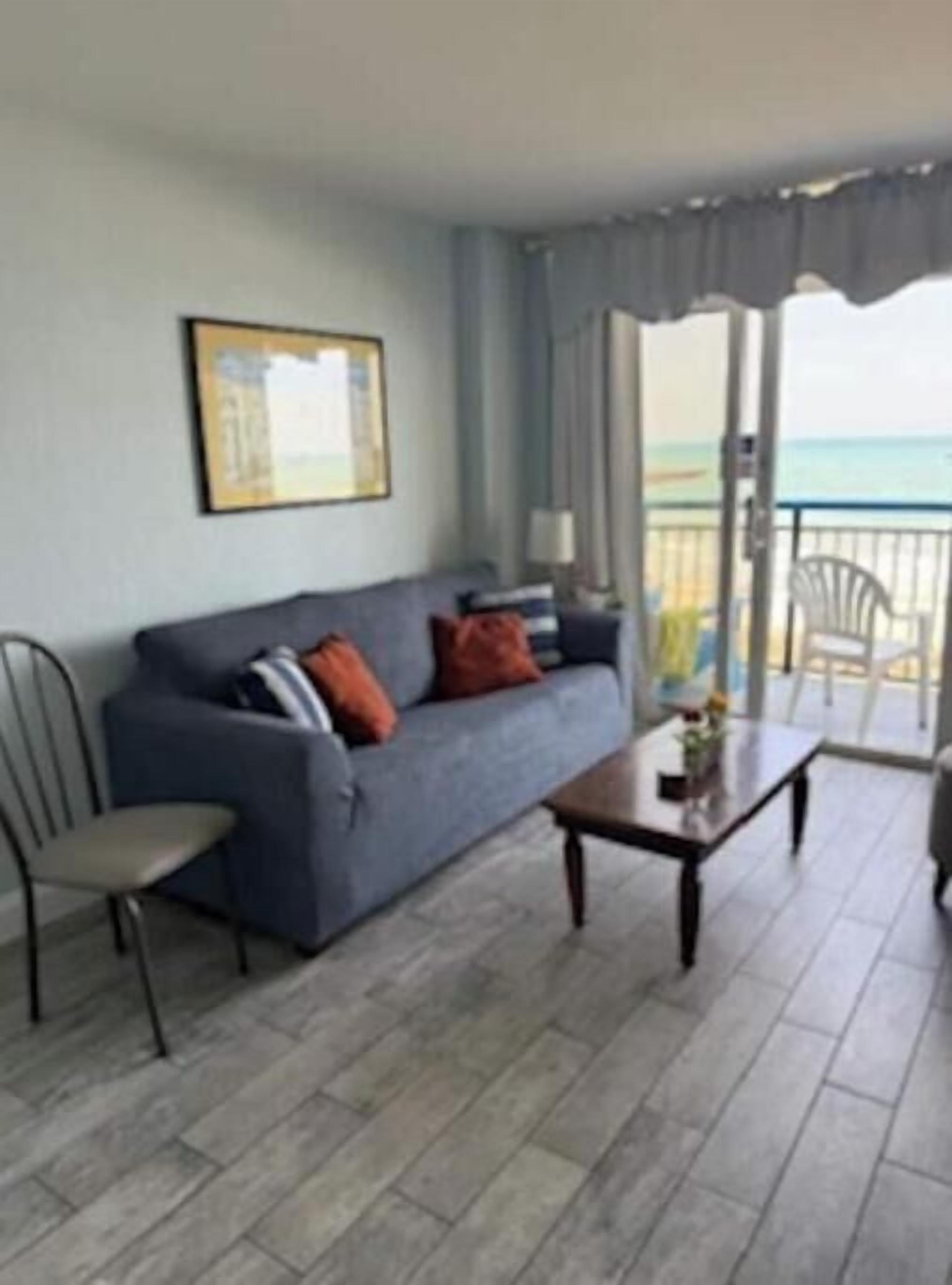 A living room with a sofa, a coffee table, and a chair. There is a balcony with a view of the ocean.