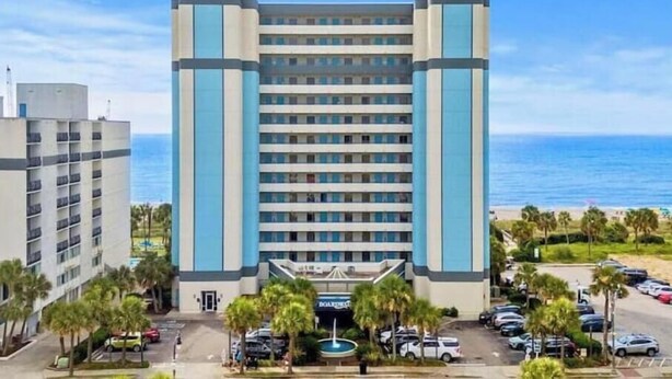 A multi-story hotel with a central entrance, parking area, and a view of the ocean. - At the heart of Myrtle Beach Oceanfront Condo (Myrtle Beach)