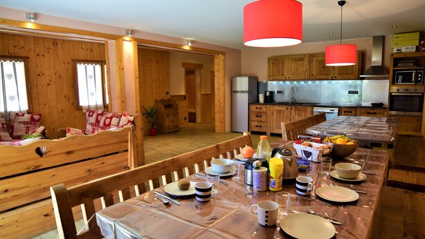 Una mesa de comedor de madera dispuesta con platos, tazas y un cuenco de fruta en una cocina con armarios de madera y una lámpara colgante roja.
