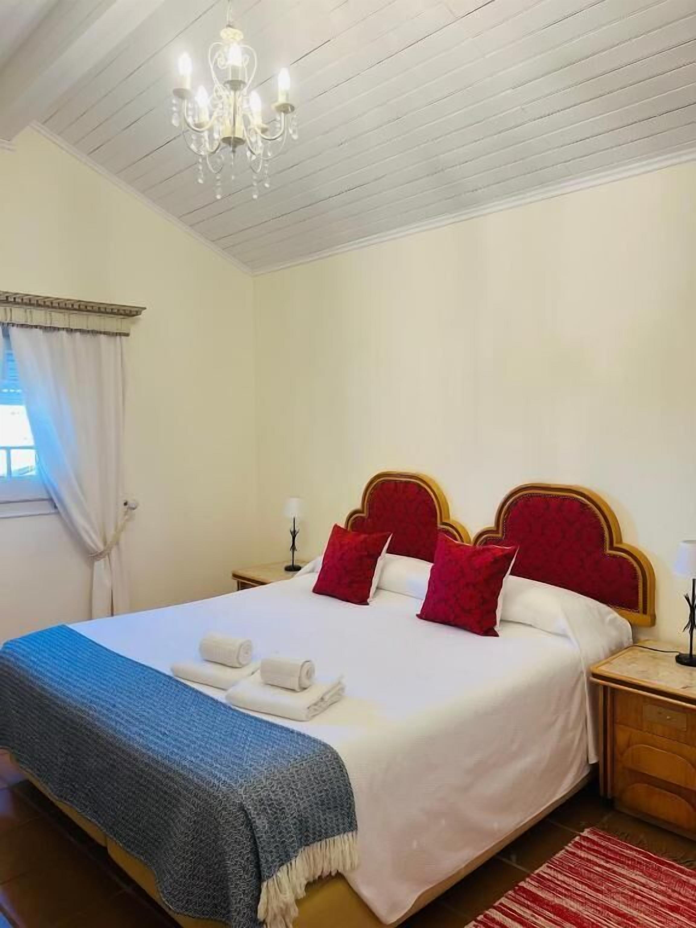 Quarto com cama, um lustre, mesa de cabeceira e janela com cortinas.