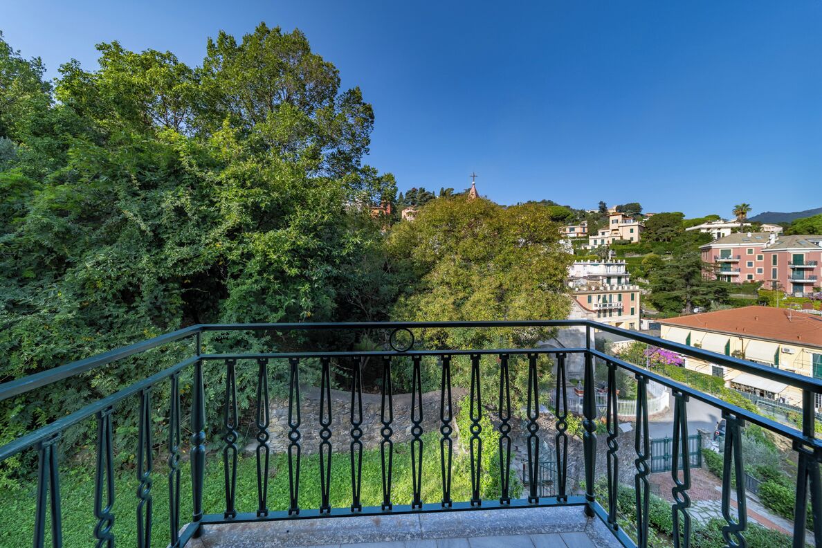 Ein Balkon mit schmiedeeisernem Geländer, der auf eine üppige, grüne Landschaft mit Bäumen und Gebäuden im Hintergrund blickt.