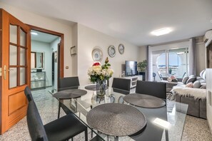 A modern living room with a glass dining table, black chairs, a sofa with a white throw, and a view of the city through a large window. - LXR Jazmines 2-Bedroom, Downtown (Fuengirola)