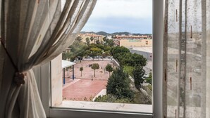 Une vue à travers une fenêtre avec des rideaux, donnant sur un terrain de basket-ball, des arbres et des bâtiments en arrière-plan.