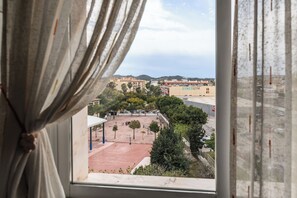 A view through a window with curtains, featuring a basketball court, trees, and buildings in the background. - LXR Jazmines 2-Bedroom, Downtown (Fuengirola)
