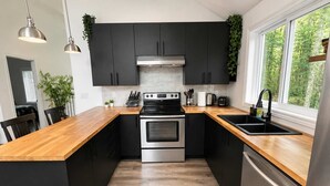 Cozinha moderna com armários pretos, eletrodomésticos de aço inoxidável, bancada de madeira e janela com vista para árvores.