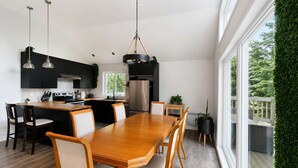 Cozinha moderna com ilha central, mesa de jantar e cadeiras, grandes janelas e vista para o verde.