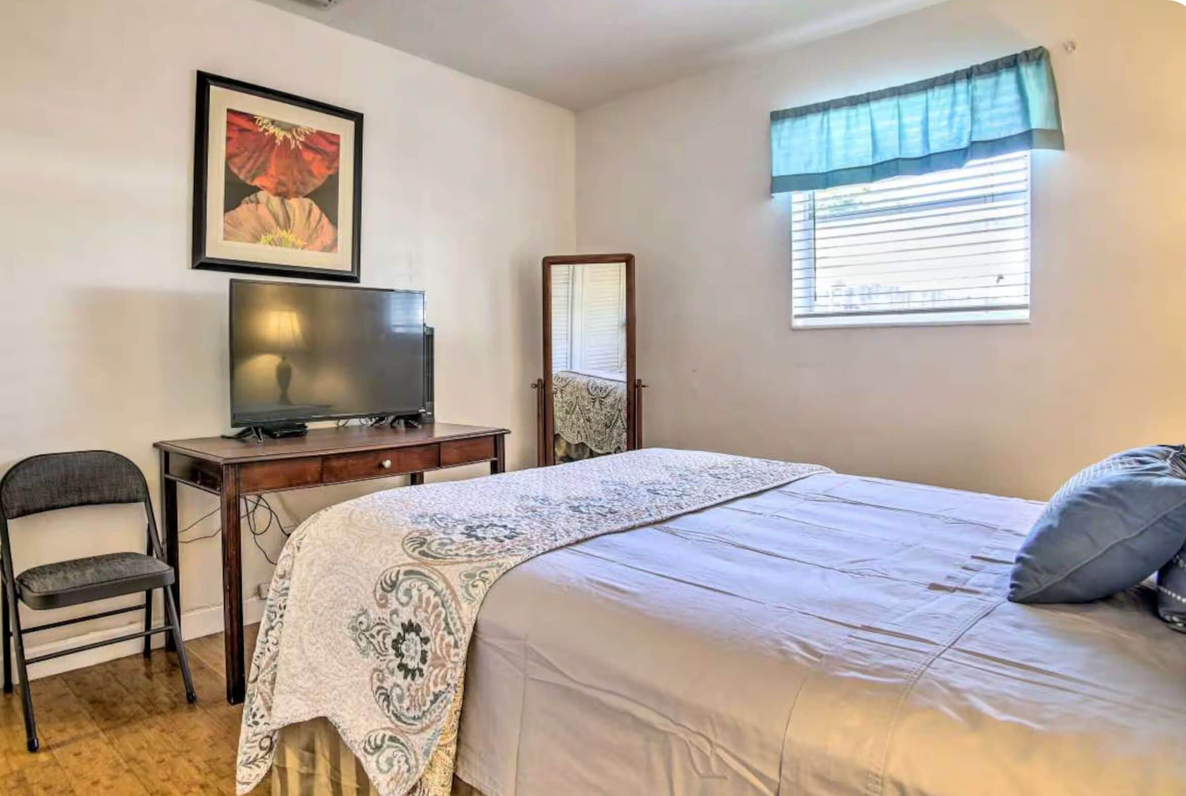 A bedroom with a bed, a desk with a television, a chair, a window with blinds, and a mirror.