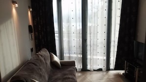 A modern living room with a large sofa, a flat-screen TV, and a view through sheer curtains.