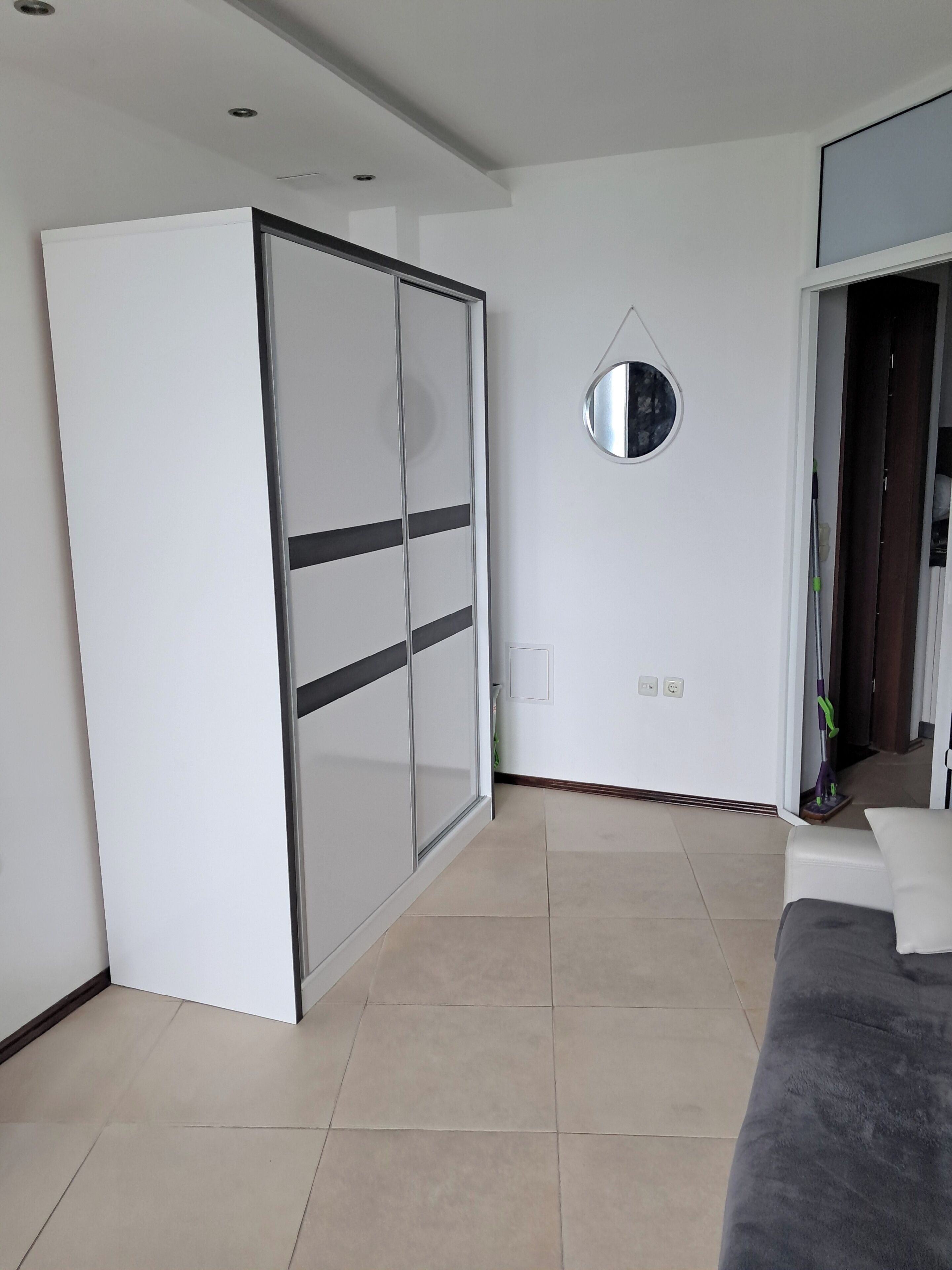 Una stanza moderna con una porta scorrevole, uno specchio rotondo, un divano grigio e un mocio appoggiato al muro.