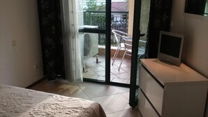 A bedroom with a bed, a TV on a bedside table, a balcony with a chair, and a sliding glass door.