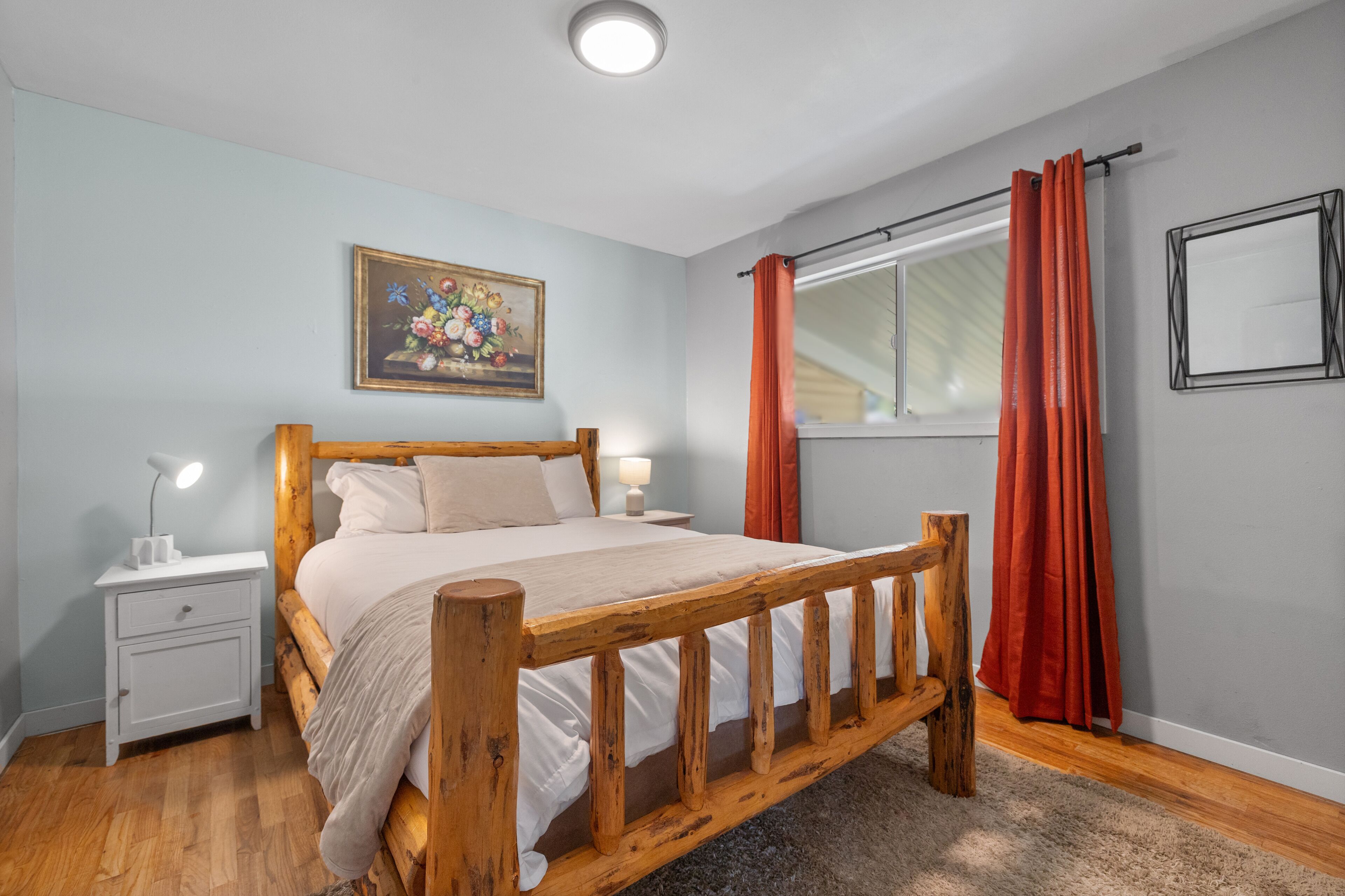 Une chambre à coucher avec un lit en bois, une table de chevet, une lampe, une fenêtre avec des rideaux et un tableau accroché au mur.