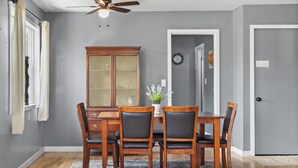 Uma sala de jantar com mesa e cadeiras de madeira, ventilador de teto, janela com cortinas, armário e porta.
