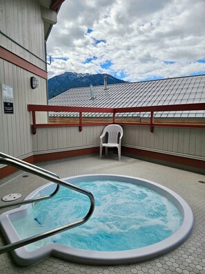 A hot tub on a deck with a view of mountains, a chair, and a safety sign. - NEW! Whistler Village Family Condo | Mountain Views + Hot Tub + Free Parking (Whistler)