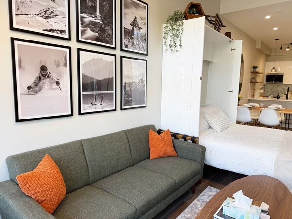 A modern living room with a grey sofa, orange cushions, a bed, a dining area, and a kitchen. - NEW! Whistler Village Family Condo | Mountain Views + Hot Tub + Free Parking (Whistler)