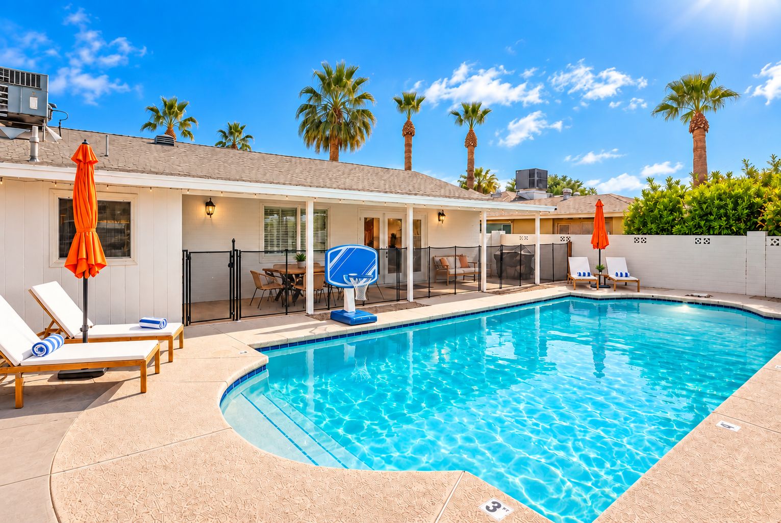 A swimming pool with lounge chairs and umbrellas, a basketball hoop, and a covered patio area.