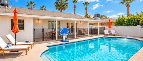 A swimming pool with lounge chairs and umbrellas, a basketball hoop, and a covered patio area.