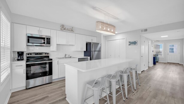 A modern kitchen with white cabinetry, a central island with bar stools, and a 'Beach' sign on the wall. - Twin Palms Beach House - Entire Duplex-Private Pool-Steps to the Beach-Sleeps 16 (Gulf Shores)