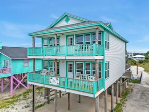 Ein zweistöckiges Strandhaus mit Balkon, einem Schild mit der Aufschrift 'Twin Palms' und einem sandigen Strand im Hintergrund.