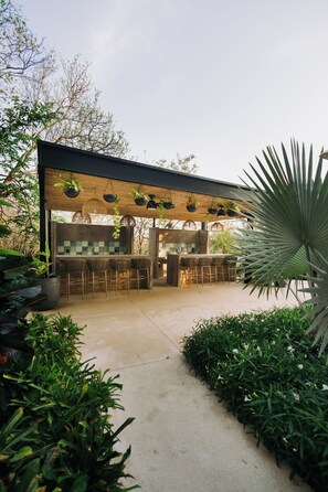 Uma área de bar ao ar livre com design moderno, incluindo balcão e assentos, cercada por vegetação e plantas.