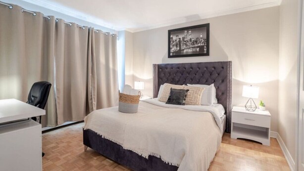 Une chambre à coucher comprenant un lit, un bureau, une chaise, une table de chevet, une lampe, un tableau au mur et une fenêtre avec des rideaux.
