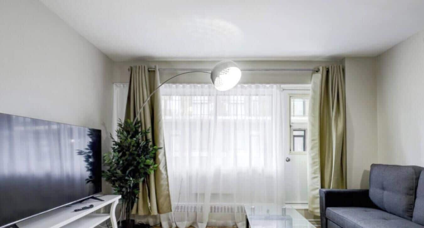 Un salón con televisor de pantalla plana, un sofá, una planta en maceta y un amplio ventanal con cortinas.