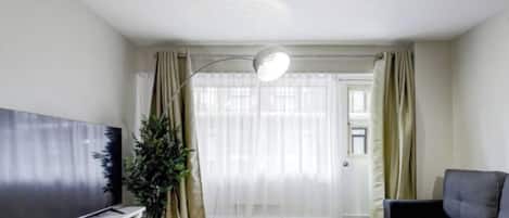 Un salón con televisor de pantalla plana, un sofá, una planta en maceta y un amplio ventanal con cortinas.