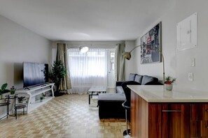 Una sala de estar moderna con televisor de pantalla plana, un sofá, una mesa de comedor y una zona de cocina con un armario de madera.