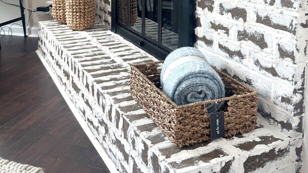 Une cheminée avec un mur en briques blanches, un téléviseur noir et deux paniers en osier posés sur un banc en bois.